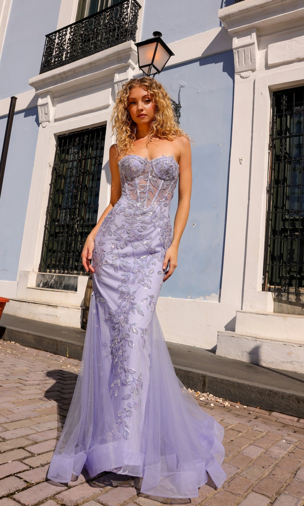 A woman wearing the Sheer-Bodice Strapless Mermaid Prom Dress G1258 with beaded embroidery stands on a cobblestone street in front of a light blue and white building.